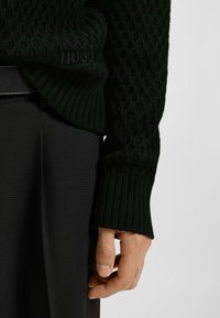 Hand mit schwarzem strukturiertem Strickärmel und geripptem Bündchen, der schwarzen Stoff in der Nähe der Hüfte hält und ein kleines Tattoo am Finger zeigt.