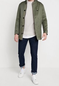 Giacca verde con chiusura a bottoni, maglione beige a trama, jeans blu navy e sneakers bianche. La giacca è dotata di colletto e tasche laterali.