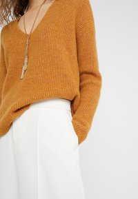 Kvinna i en texturerad orange V-ringad tröja instoppad i högmidjade vita byxor med en hand i fickan och en gyllenfransad halsband.