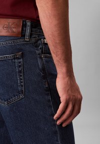 Tumed sinised teksapüksid, millel on nahast Calvin Kleini plaaster, nähtavad õmblused ja vormis lõige, kus üks käsi toetub vöökoha peal.