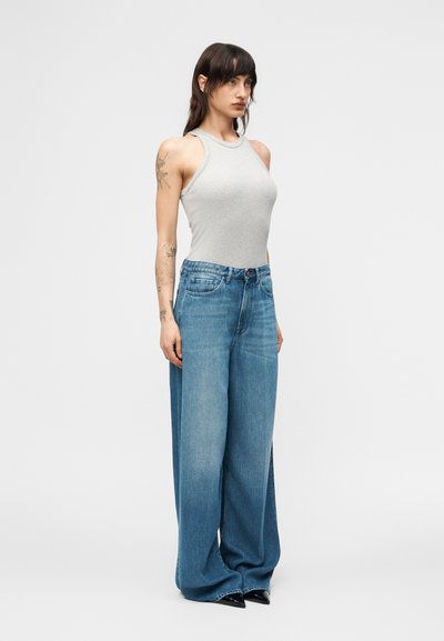 Γυναίκα με μαλλιά μέχρι τον ώμο, που φοράει στενή γ κρι αμάνικη μπλούζα, μπλε τζιν παντελόνι με φαρδιές παντελόνες και μαύρα μυτερά παπούτσια, που στέκεται πλαγίως.