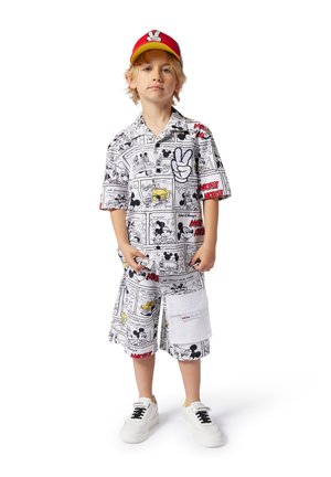 Chemise blanche à manches courtes et shorts avec un imprimé de bande dessinée noir de Mickey Mouse ; poche blanche sur les shorts ; casquette rouge et jaune ; baskets blanches.