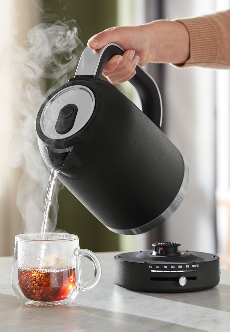 Czarny elektryczny czajnik nalewający parę i wodę do przezroczystej szklanej filiżanki wypełnionej herbatą, stojącej na ciemnej podstawie z pokrętłem temperatury.