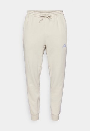 Beige joggebukser med elastisk midje, snøring, smalnende ankler, og lite logo på høyre lår mot en enkel hvit bakgrunn.