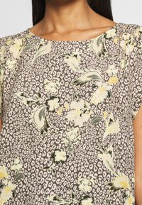 Blus med blommönster i ljusgrått med gula och svarta accenter; har rundringning och korta ärmar med diskret samlingsdetalj.