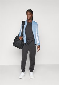 Puma treningsdrakt med en lyseblå jakke og svarte bukser, med hvite striper, kombinert med en svart treningsbag og hvite joggesko.