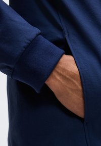 Χέρι που ξεκουράζεται μέσα στην τσέπη ενός σκούρου μπλε μπουφάν με ριμπ τελείωμα στο μανίκι.