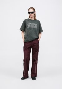 Zaļš, plankumains, pārāk liels Nike T-krekls ar baltu logo, apvienots ar tumši sarkaniem, brīvi piegulošiem biksēm ar baltām sānu svītrām un melniem saulesbrillēm.