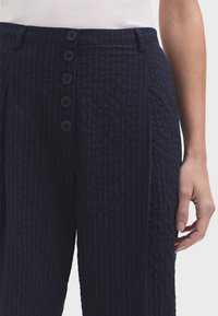Tmavoslijepe hlače s teksturom koje sadrže vertikalne pruge, zatvaranje na gumbe s šest tamnih gumba i nabranim dizajnom za dodatnu dimenziju.