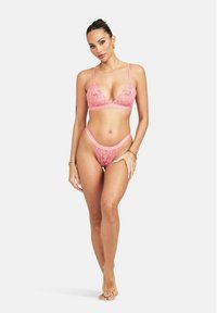 Een model poseert in een set van roze kanten lingerie, bestaande uit een bralette en bijpassende string, met een neutrale achtergrond die de outfit benadrukt.