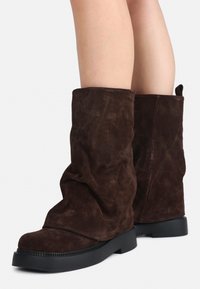 Stivali in suede marrone a metà polpaccio con suole in gomma nera indossati su gambe nude, mostrano lievi pieghe sulla parte superiore degli stivali.