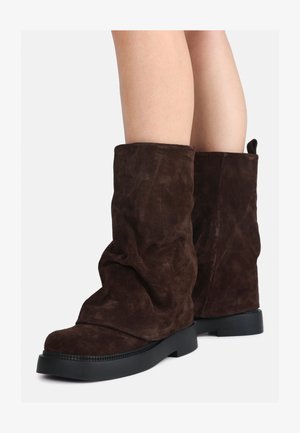 Brown suede mid-calf boots with black rubber soles worn on bare legs, showing slight wrinkling on the boot shafts.