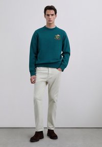 Hombre de pie vistiendo sudadera azul verdosa con gráfico de montaña, pantalones beige claro y botas marrón oscuro frente a pared clara y lisa.