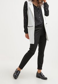 Blazer blanc sans manches porté sur un chemisier noir à motifs, associé à un pantalon noir fuselé et des mocassins noirs massifs. Tissus texturés, design minimaliste.