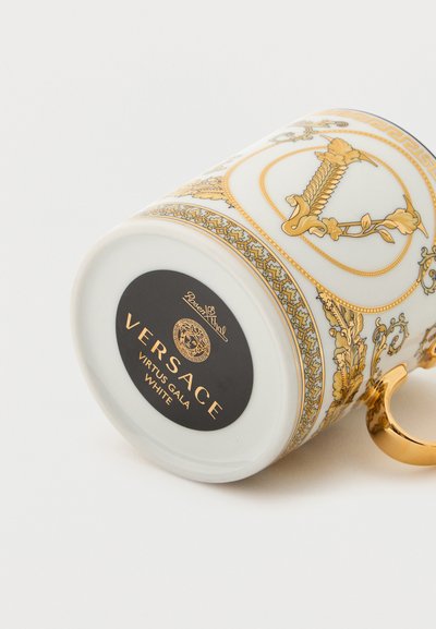 Λευκή πορσελάνινη κούπα Versace Virtus Gala με περίτεχνα χρυσά ανθικά μοτίβα και λαμπερή χρυσή λαβή, απεικονιζόμενη ξαπλωμένη στο πλάι της.