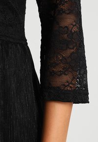 Musta mekko, jossa on pitsinen yläosa kukkakuosisella ja pitkät pitsihihat. Hame on taiteltu, mikä lisää tekstuuria ja ulottuvuutta.