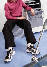 Zwarte wijde broek met een roze hoodie, gecombineerd met zwarte en zilveren sneakers. Op de achtergrond is een printer te zien en een blauwe vloer.