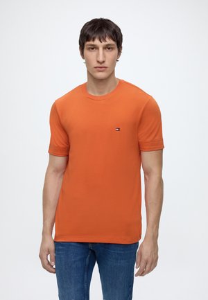 Jeune homme aux cheveux courts et foncés, portant une chemise orange à manches courtes et un jean bleu, debout devant un fond blanc uni.