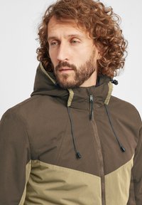 Brown and olive green hooded jacket with a zip closure, featuring a geometric color block design and drawstring accents on the hood.