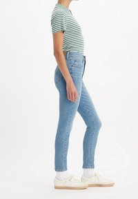Camisa corta de rayas verdes y blancas, pantalones vaqueros ajustados de talle alto en azul claro y zapatillas blancas con suelas de goma, visto de lado.