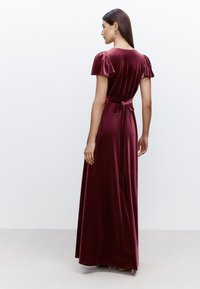 Robe maxi en velours bordeaux avec manches bouffantes, corsage ajusté et détail noué au dos. Texture lisse et longueur jusqu'au sol.