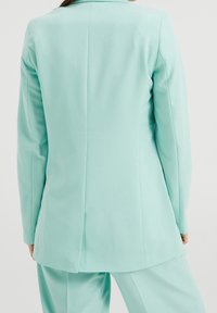 Mátově zelený tailored blazer s hladkou texturou, jedním zadním rozparkem a dlouhými rukávy. Design je minimalistický, bez viditelného hardwaru.