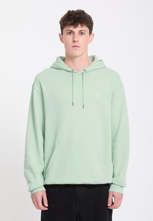 Jeune homme aux cheveux courts et bouclés porte un sweat à capuche vert clair avec poche avant et un pantalon noir, debout devant un fond blanc.