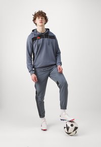 Grauer Sportanzug mit schwarzen Akzenten, bestehend aus einem mit Reißverschluss versehenen Kapuzenoberteil und tapered Hosen, kombiniert mit einem schwarz-weißen Fußball.