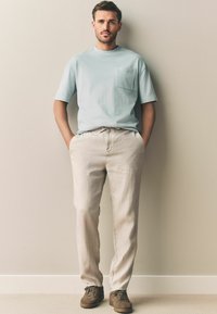 Lyseblå T-shirt med korte ærmer og lomme, parret med beige linnedbukser og brune ruskindsko. Enkel, afslappet silhuet.