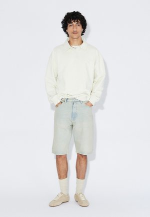 Jasnobeżowa długa koszulka polo z długim rękawem, niebieskie krótkie spodenki dżinsowe, beżowe tenisówki i białe skarpetki. Model stoi z rękami w kieszeniach na jednolitym tle.