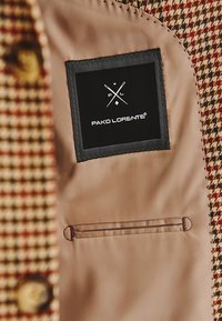 Materiał w brązową kratkę houndstooth z beżową podszewką, z czarną metką z białym tekstem i logo oraz małą wewnętrzną kieszonką.