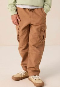 Kind met koffiebruin cargobroek met klepzakken en beige sneakers, handen in de zakken, staand op een lichte vloer tegen een effen achtergrond.