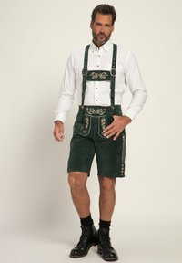 Grønne suede lederhosen med broderede detaljer, justerbare stropper og knapper på siden, parret med en hvid skjorte med knapper og sorte støvler.