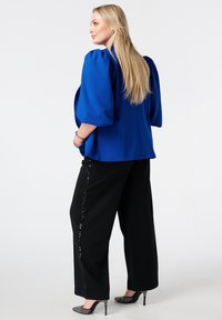Blauwe blazer met pofmouwen en zwarte wijde broek met paillettenaccenten aan de zijkant. Model draagt grijze, puntige hakken, wat een slanke silhouet benadrukt.