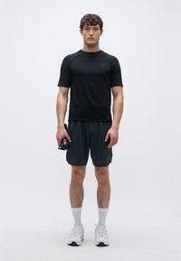 Tricou sport negru cu mânecă scurtă asortat cu pantaloni scurți închiși la culoare. Modelul ține o sticlă de apă neagră, purtând încălțăminte sport albă și șosete.