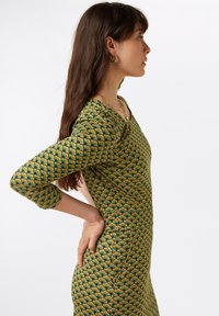 Pitkähihainen, istuva mekko, jossa on vihreä ja keltainen geometrinen kuvio, V-muotoinen kaulus ja teksturoitu kangas.