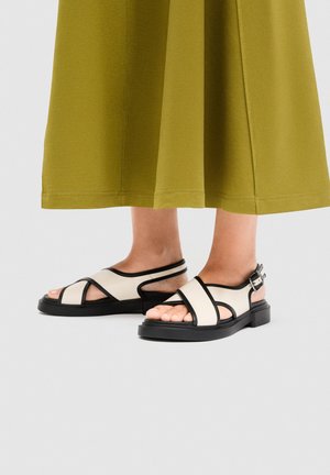 Picioare purtând sandale crem cu margini și catarame negre, asortate cu o fustă verde măsliniu care se termină la jumătatea gambei, pe un fundal gri deschis.