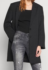 Černý oversized blazer s klopami, dvěma knoflíky a dlouhými rukávy, vrstvený přes přiléhavý černý top a potrhané šedé džíny.
