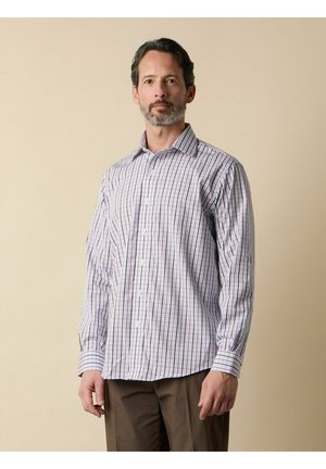 Mężczyzna o krótkich ciemnych włosach i brodzie, ubrany w białą koszulę w kratę z długim rękawem i brązowe spodnie, stojący na beżowym tle.