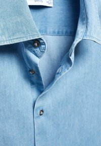 Chemise en denim bleu clair avec un col pointu, dotée de boutons marron et d'une texture douce, mettant en valeur les détails de couture et de tissu.