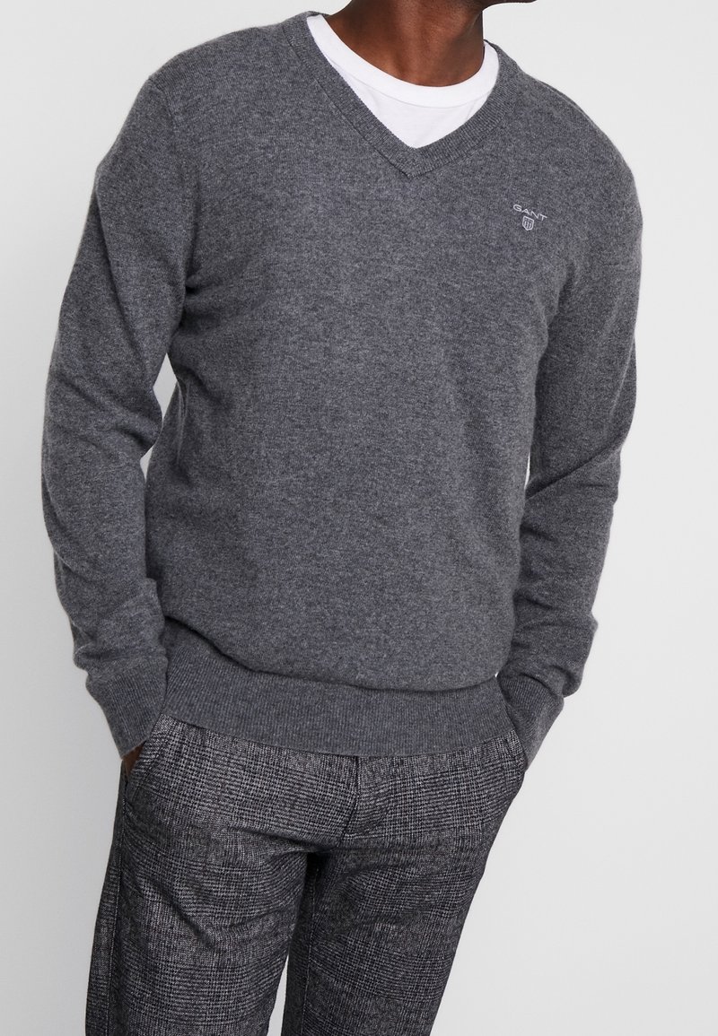 Homme portant un pull gris en V sur une chemise blanche, avec un pantalon à carreaux gris, les mains dans les poches, sur un fond uni.