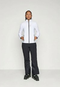 Weiße Zip-Jacke mit Seitentaschen, kombiniert mit schwarzen Skihosen und schwarzen Skischuhen. Glattes Material und taillierter Schnitt.