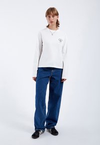 White long-sleeve t-shirt with a heart graphic, paired with loose dark blue denim jeans and black athletic sneakers. Smooth fabric texture.