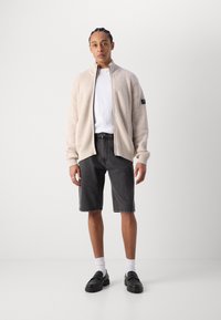 Beige ribbad zip-up kofta över en vit t-shirt, matchad med svarta jeansshorts och svarta loafers. En enkel design med kontrasterande texturer.