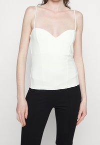 Femme portant un haut blanc ajusté sans manches avec fines bretelles et un pantalon noir, debout les bras détendus le long du corps.