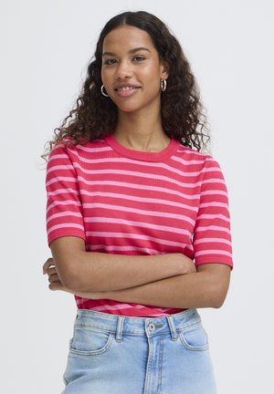 Jonge vrouw met krullend haar, gekleed in een roze en rood gestreept shirt en lichtblauwe spijkerbroek, met gekruiste armen, licht glimlachend, neutrale achtergrond.