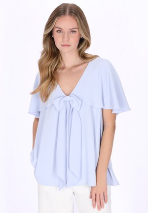 Kobieta z długimi falującymi włosami nosi jasnoniebieską bluzkę z dekoltem w kształcie litery V, krótkimi, rozkloszowanymi rękawami i kokardą z przodu, w połączeniu z białymi spodniami.