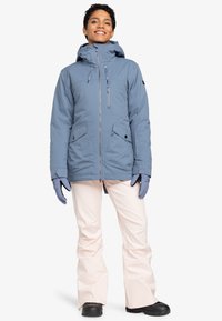 Light blue insulated jacket with hood, two front pockets, zippers, paired with soft pink snow pants and black shoes. Hands covered in gloves.
