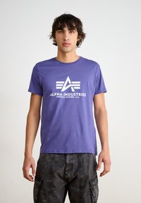 Violets kokvilnas T-krekls ar īsām piedurknēm, ar baltu logo un tekstu "ALPHA INDUSTRIES" centrā priekšpusē, ar apaļu kakla izgriezumu.