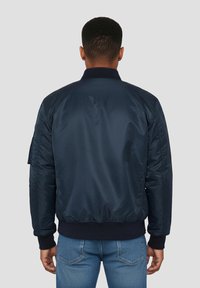 Veste bombardier marine avec une texture lisse et brillante ; dotée de poignets et d'un ourlet côtelés ; sans marque ni motif visible ; portée avec un jean bleu.